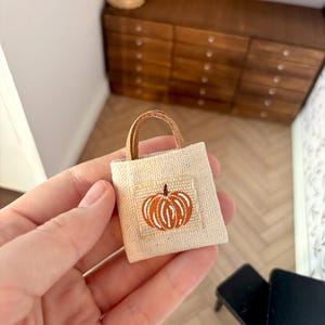 Op de afbeelding: Een miniatuur beige draagtas met een gestikt oranje pompoenontwerp. De tas heeft een kleine, rechthoekige patch met de pompoen en een bruin handvat. De tas wordt in een hand gehouden, wat duidt op een poppenhuisaccessoire.