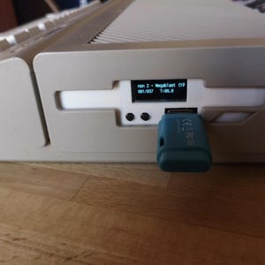 May include: A beige vintage computer console featuring a small digital display screen showing the text "non 2 - Megablast (19 881/837 T:00.0". A teal USB flash drive is inserted into the front port, positioned next to two small black circular buttons.