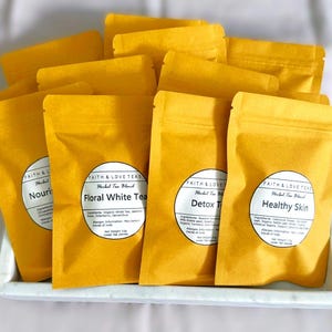 May include: A collection of yellow herbal tea pouches, each with a white label. The labels read "Nourish", "Floral White Tea", "Detox Tea", and "Healthy Skin". The pouches are arranged on a white tray.