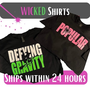 May include: Two black t-shirts with graphic designs. One shirt says "Defying Gravity" in white and green, with a witch silhouette. The other says "Popular" in pink. The top banner reads "Wicked Shirts".