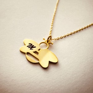 May include: A gold necklace with a bee-shaped pendant. The bee has a cutout design and the letter "M" is visible inside the bee's body.