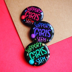 May include: Three black button pins with the text "Support Girls in STEM" in colorful lettering. The buttons are arranged in a stack on a brown and red background.