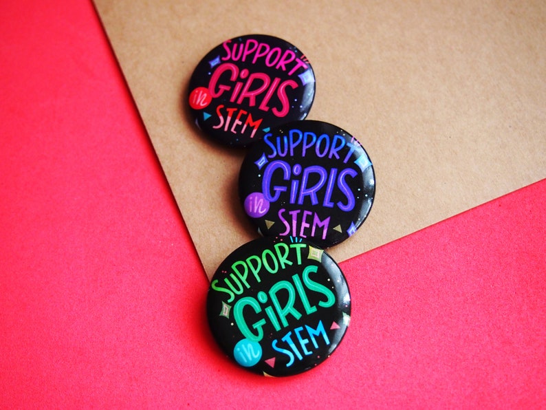 May include: Three black button pins with the text "Support Girls in STEM" in colorful lettering. The buttons are arranged in a stack on a brown and red background.