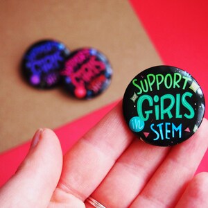 May include: A black button with glitter and the text "SUPPORT GIRLS in STEM" in green and white lettering.