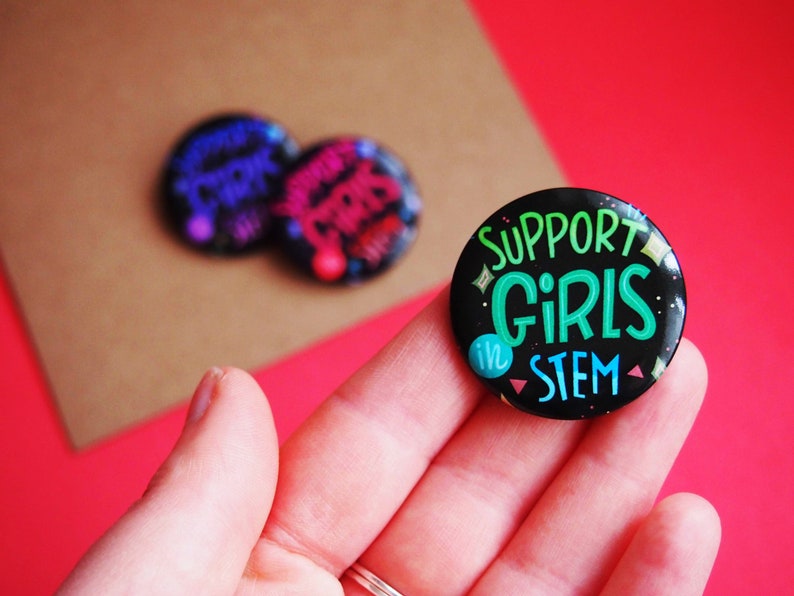 May include: A black button with glitter and the text "SUPPORT GIRLS in STEM" in green and white lettering.