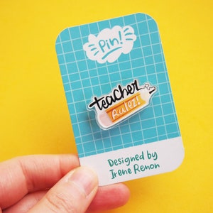 Puede incluir: Un pin de esmalte en forma de lápiz amarillo y blanco con el texto "teacher rulez!" y la palabra "Pin!" en una nube blanca. El pin está sobre un fondo de cuadrícula azul y blanco con el texto "Designed by Irene Renon".