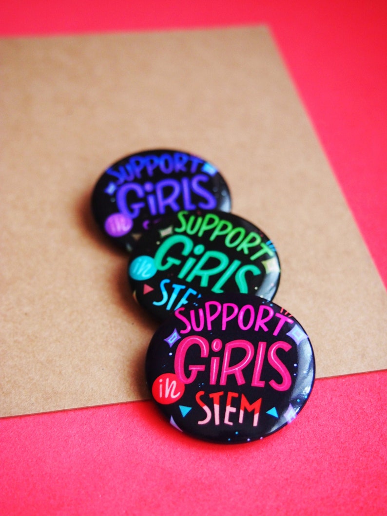 May include: Three black button pins with the text "Support Girls in STEM" in different colors. The top pin has purple text, the middle pin has green text, and the bottom pin has pink text.