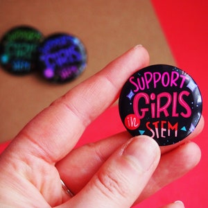 May include: A black button with a pink and blue design that says "Support Girls in STEM".