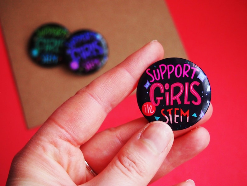 May include: A black button with a pink and blue design that says "Support Girls in STEM".