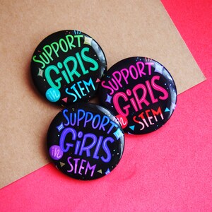 May include: Three black button pins with the text "Support Girls in STEM" in colorful lettering. The buttons are on a brown and pink background.