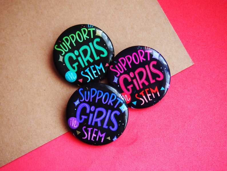 May include: Three black button pins with the text "Support Girls in STEM" in colorful lettering. The buttons are on a brown and pink background.