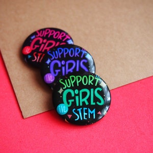 May include: Three black button pins with the text "Support Girls in STEM" in different colors. The pins are stacked on top of each other on a brown surface.