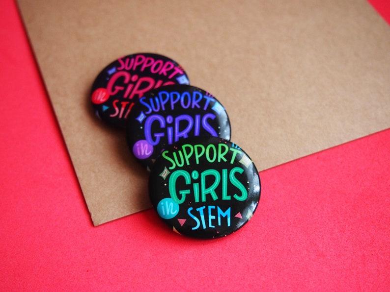 May include: Three black button pins with the text "Support Girls in STEM" in different colors. The pins are stacked on top of each other on a brown surface.