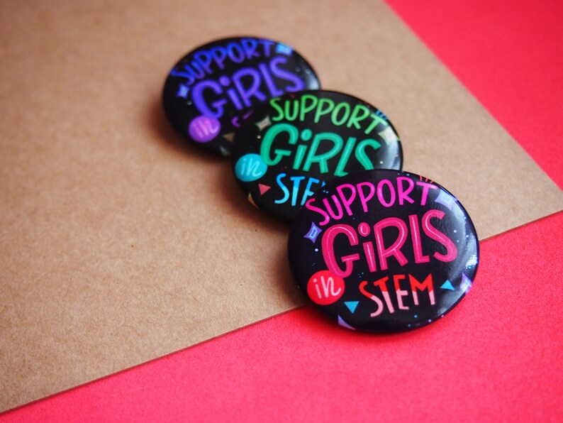 May include: Three round black button pins with a white background and colorful text. The text on the pins reads "Support Girls in STEM".