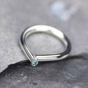 Op de afbeelding: Een zilveren ring met een V-vormige band en twee kleine edelstenen. De ring heeft een blauwe edelsteen aan de ene kant en een heldere edelsteen aan de andere.
