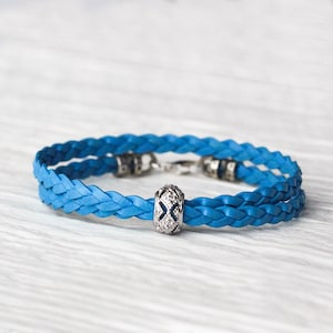 May include: A blue braided leather bracelet with a silver charm featuring a decorative design and small clear stones.