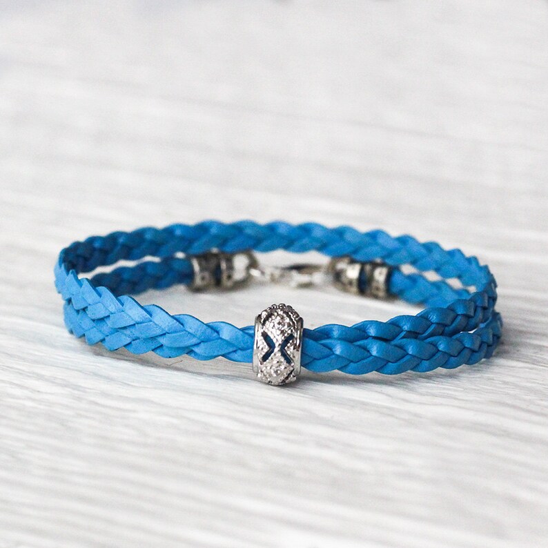 May include: A blue braided leather bracelet with a silver charm featuring a decorative design and small clear stones.
