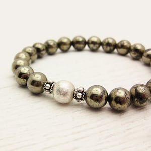 May include: A bracelet made of dark brown pyrite beads with a silver bead in the centre. The bracelet is on a white surface.
