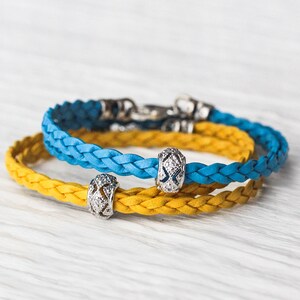 May include: Two braided leather bracelets, one blue and one yellow, with silver charm beads. The bracelets are stacked on top of each other.