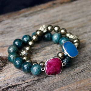 Puede incluir: Un brazalete de doble cadena con cuentas de piedras preciosas azules y rojas. El brazalete está hecho con cuentas de apatita azul, rubí rojo y pirita dorada.