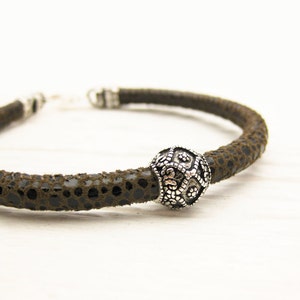 May include: A brown leather bracelet with a silver bead featuring a decorative design. The bracelet is made of a textured leather material.