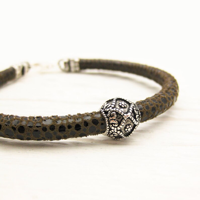 May include: A brown leather bracelet with a silver bead featuring a decorative design. The bracelet is made of a textured leather material.