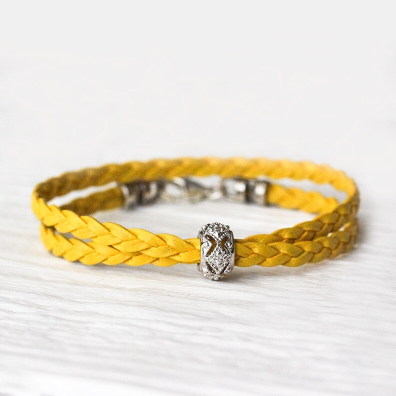 May include: A yellow braided leather bracelet with a silver charm featuring a geometric design with clear rhinestones.