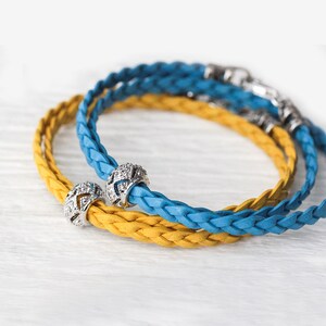 May include: Two braided leather bracelets, one yellow and one blue, with silver bead charms. The charms are decorated with small, clear crystals.