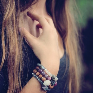 Op de afbeelding: Een vrouw draagt een zwart shirt met meerdere armbanden om haar pols. De armbanden zijn gemaakt van hout, kralen en metaal. De armbanden zijn gelaagd en hebben verschillende kleuren en texturen.