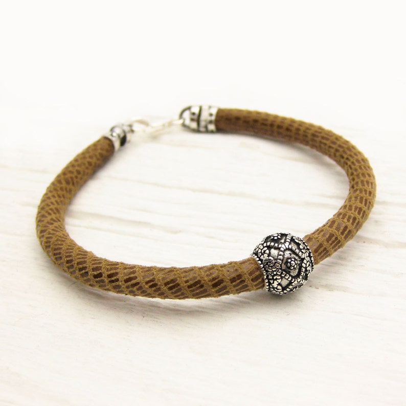 May include: A brown leather bracelet with a silver bead featuring a decorative design. The bracelet has a clasp closure.
