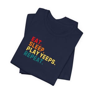 May include: A navy blue t-shirt with the words "EAT. SLEEP. PLAY YEEPS. REPEAT." in a retro, multi-colored font. The shirt is folded and appears to be made of soft material.
