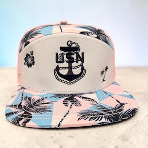 Könnte beinhalten: Eine Baseballkappe mit einem hellrosa und blauen geometrischen Muster und schwarzen Palmensilhouetten. Das Vorderteil ist cremeweiß und zeigt ein schwarzes, gesticktes Ankerlogo mit den Buchstaben USN, die mittig im Anker platziert sind.