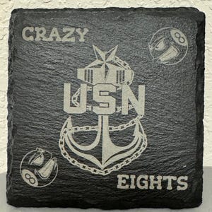 May include: A black slate coaster with a white engraved anchor, chain, and the text "CRAZY EIGHTS USN". There are two white engraved 8-ball images on the coaster.