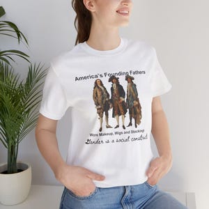 May include: White t-shirt with a graphic of three men in colonial attire. The text reads "America's Founding Fathers Wore Makeup, Wigs and Stockings Gender is a social construct."