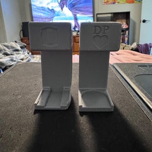 May include: Two gray 3D printed dice towers, one with the letter "D" and the other with the letters "DP" and a heart symbol.