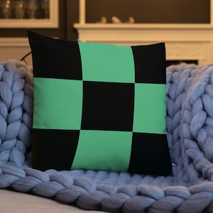 May include: A square decorative pillow with a black and mint green checkered pattern. The pillow is resting on a light gray knitted blanket and a pink sofa. The pillow is a decorative home accent.