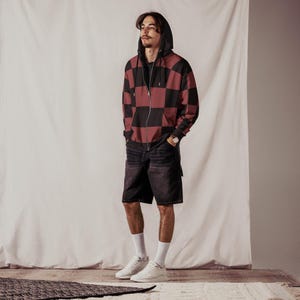 Puede incluir: Una persona lleva una sudadera con capucha de cremallera a cuadros negros y burdeos con una sudadera con capucha negra debajo. Tambi&eacute;n lleva pantalones cortos de mezclilla oscura y zapatillas blancas.
