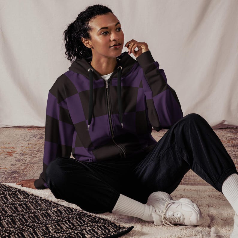 Puede incluir: Una sudadera con capucha de cremallera negra y p&uacute;rpura a cuadros. La modelo lleva la sudadera y pantalones negros.