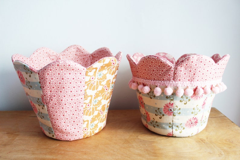 Peut inclure: Deux paniers de rangement en tissu avec des bords festonn&eacute;s. Le plus grand panier est rose avec des pois blancs et un motif floral. Le plus petit panier est rose avec des pois blancs et un motif ray&eacute; avec des fleurs roses. Les deux paniers ont une bordure en pompons roses.