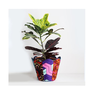 May include: A colourful, patterned fabric planter with a scalloped rim, holding a green and red leafy plant. The planter is made of fabric and has a patchwork design with geometric shapes and bright colours.