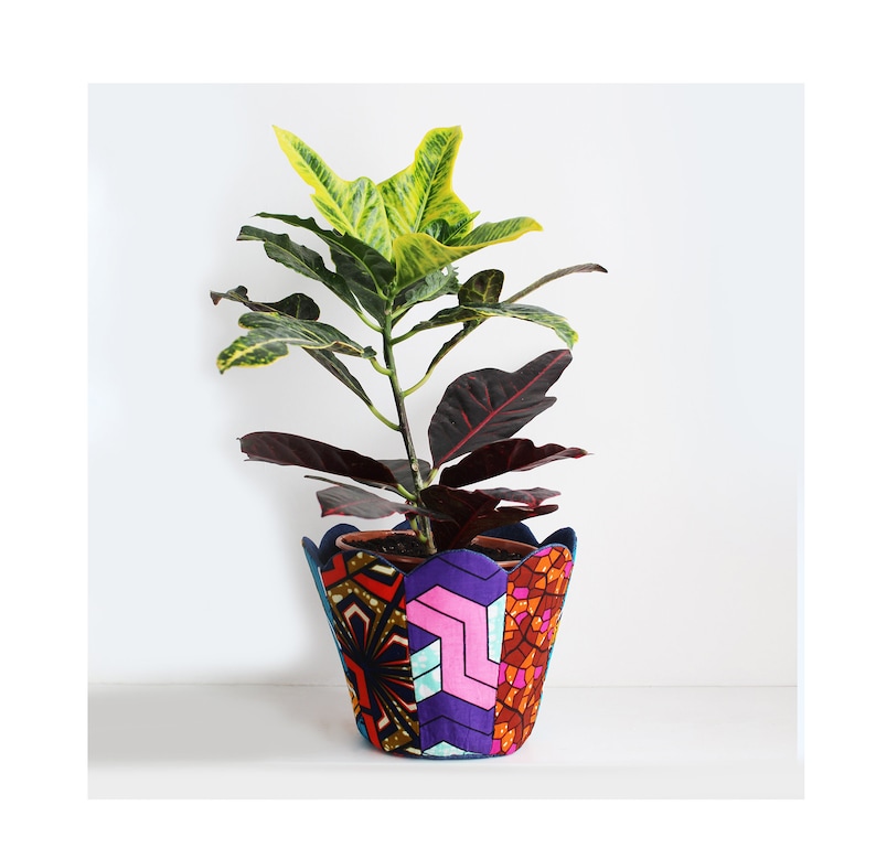 Peut inclure: Un pot de fleurs en tissu color&eacute; et &agrave; motifs, avec un bord festonn&eacute;, contenant une plante verte et rouge. Le pot est fait de tissu et pr&eacute;sente un motif patchwork avec des formes g&eacute;om&eacute;triques et des couleurs vives.