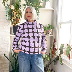 May include: A long-sleeved turtleneck shirt featuring a repeating pattern of large, light purple circles on a dark brown background. The shirt is paired with blue denim jeans. The person is standing in front of a window with plants.