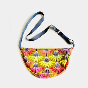 May include: A crescent-shaped crossbody bag featuring a quilted olive green fabric with a repeating pattern of vibrant pink, red, and yellow coneflower blossoms. The bag includes a solid dark blue adjustable nylon strap with a metal slider.