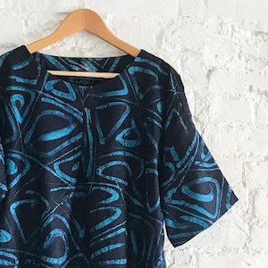 May include: A short-sleeved top featuring a dark black base color with a repeating abstract pattern in bright cyan blue. The garment has a wide, rounded neckline and is displayed on a wooden hanger, showcasing the fabric's intricate batik-style design.