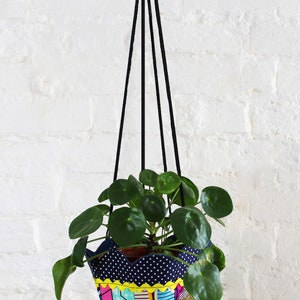 Peut inclure: Un pot de fleurs suspendu en tissu color&eacute; avec un cordon noir, contenant une plante verte aux feuilles rondes. Le pot a un motif g&eacute;om&eacute;trique bleu, jaune, rose et blanc.