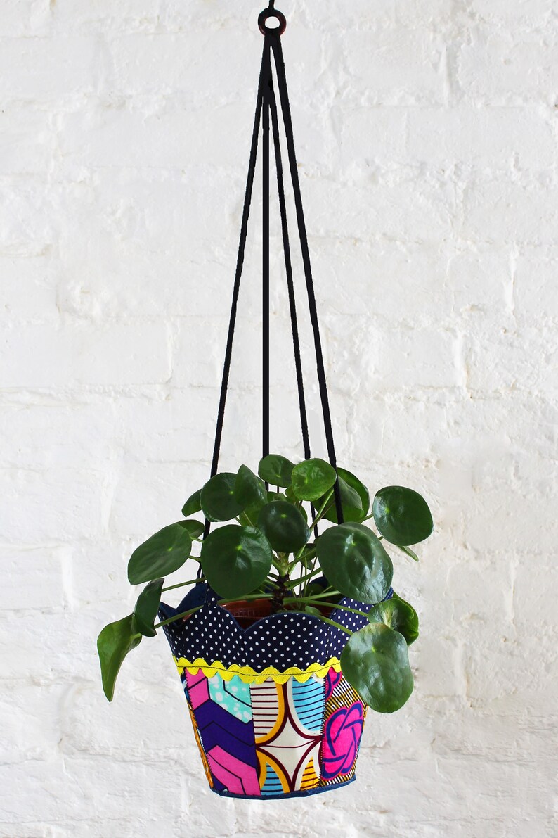 Peut inclure: Un pot de fleurs suspendu en tissu color&eacute; avec un cordon noir, contenant une plante verte aux feuilles rondes. Le pot a un motif g&eacute;om&eacute;trique bleu, jaune, rose et blanc.