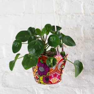 Peut inclure: Un pot suspendu en tissu color&eacute; &agrave; motifs floraux. Le pot est rempli d'une plante verte aux feuilles rondes.