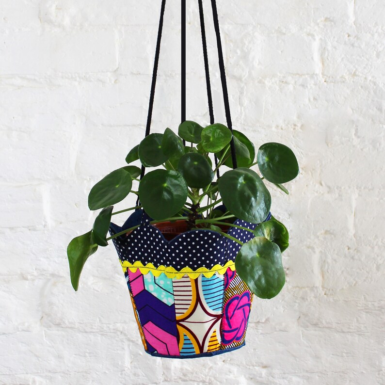 Peut inclure: Un pot suspendu en tissu avec un motif &agrave; pois bleus et blancs, une bordure jaune et un motif g&eacute;om&eacute;trique color&eacute;. Le pot est rempli d'une plante verte aux feuilles rondes.