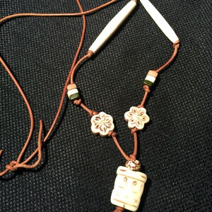 May include: A brown leather cord necklace with white bone beads, including two long cylindrical beads, two flower-shaped beads, and a rectangular pendant with a carved design.