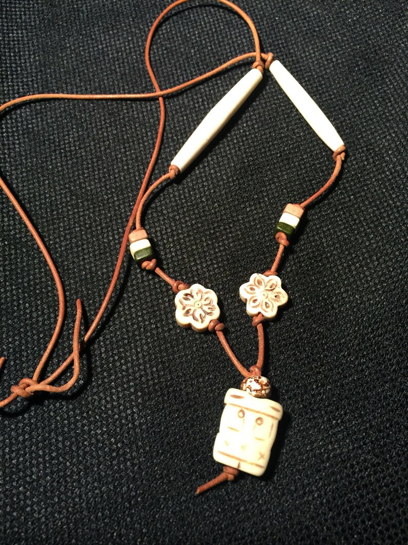 May include: A brown leather cord necklace with white bone beads, including two long cylindrical beads, two flower-shaped beads, and a rectangular pendant with a carved design.